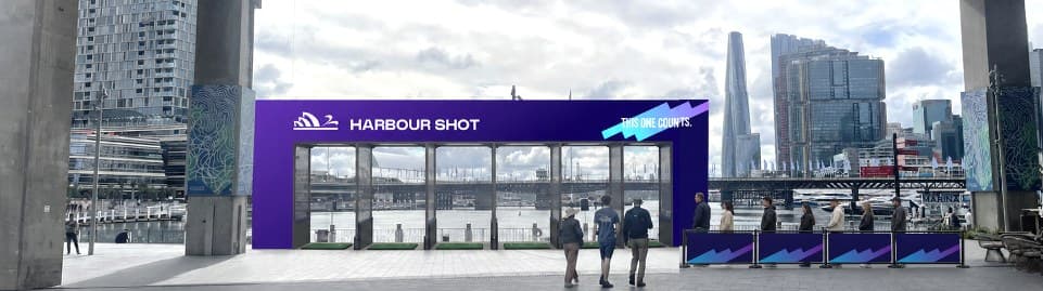 Artist's impression: a purple Harbour Shot enclosure with six tee bays on the Cockle Bay promenade, facing the water, with the Barangaroo skyline behind and people queuing at branded barriers.